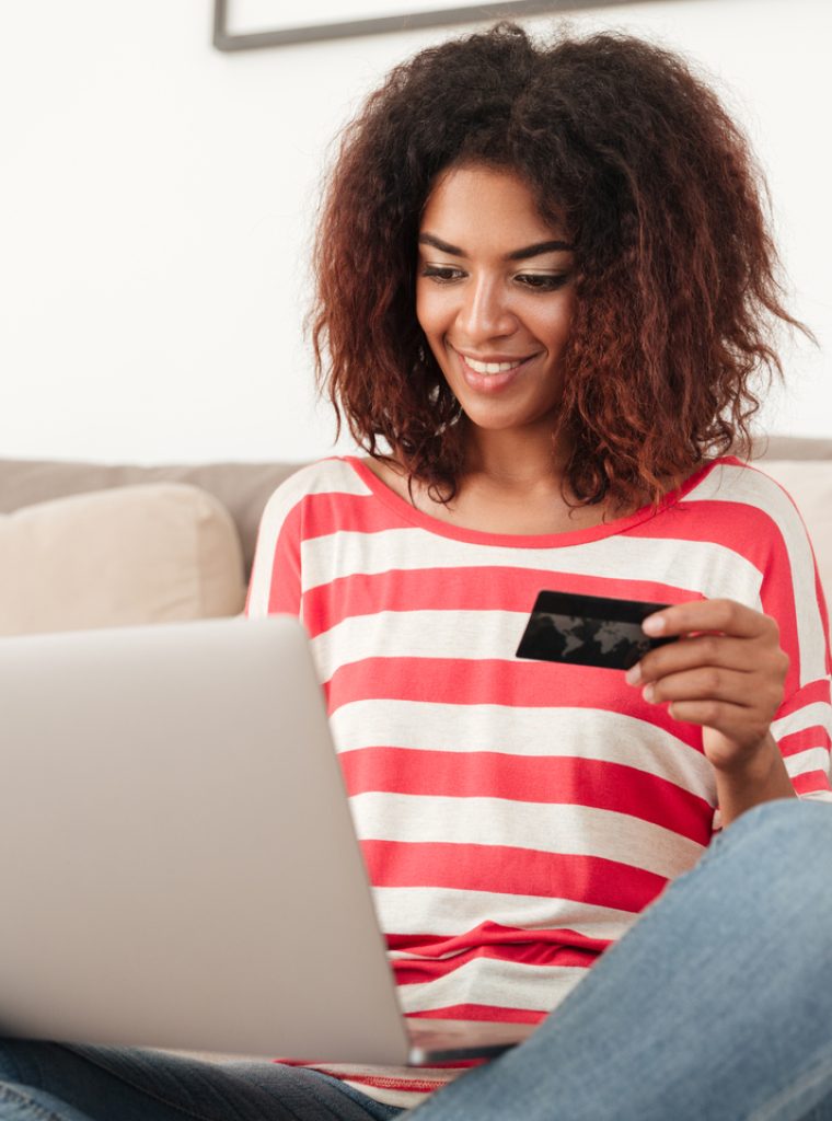 Smiling african woman sitting on bed with credit card and laptop computer at home